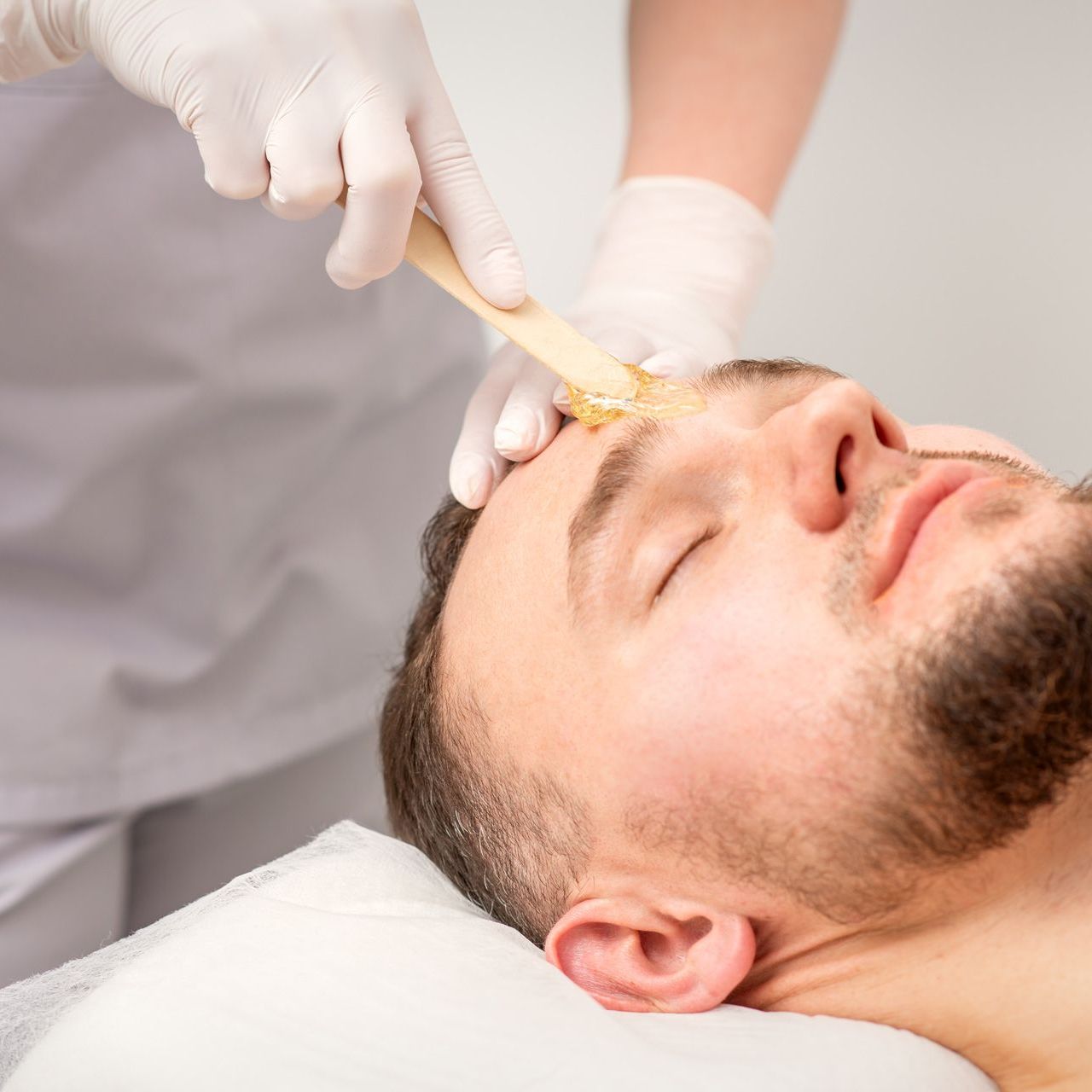 Gloved hands applying wax to a man’s eyebrow during a beauty treatment