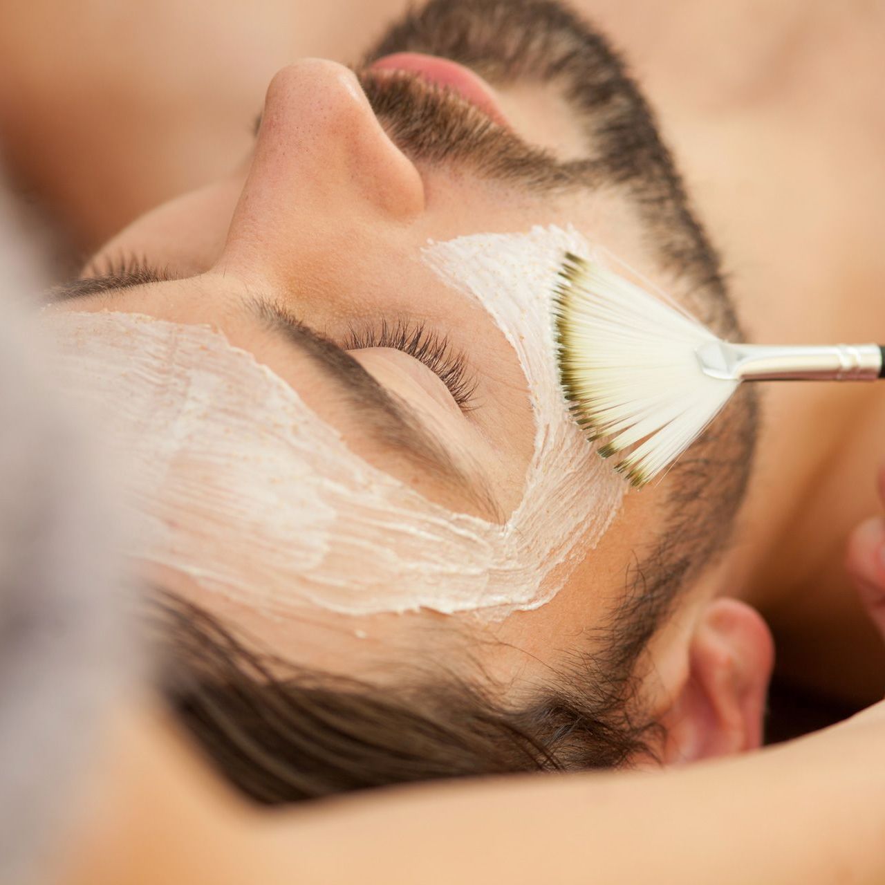 Spa treatment with a brush applying white cream to a man's forehead and face